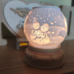 Puede incluir: Un portavelas redondo de cerámica blanca con una escena invernal con muñecos de nieve. El portavelas se asienta sobre una base marrón y tiene un brillo cálido de la luz interior. El diseño incluye nieve cayendo.