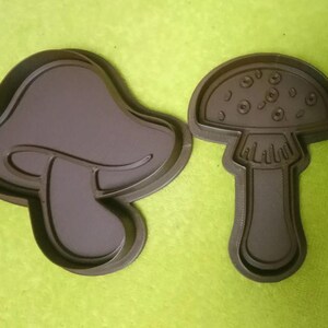May include: Two dark brown mushroom-shaped cookie cutters. One is a classic mushroom shape, and the other has a more abstract design. The cutters are made of plastic and are set against a green background.