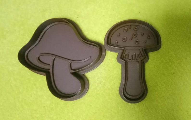 May include: Two dark brown mushroom-shaped cookie cutters. One is a classic mushroom shape, and the other has a more abstract design. The cutters are made of plastic and are set against a green background.