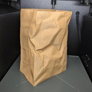 May include: A crumpled brown paper bag stands upright on a gray surface, likely inside a 3D printer. The bag has a rectangular base and an open top, with visible creases and folds throughout its surface. The printer's components are visible in the background.