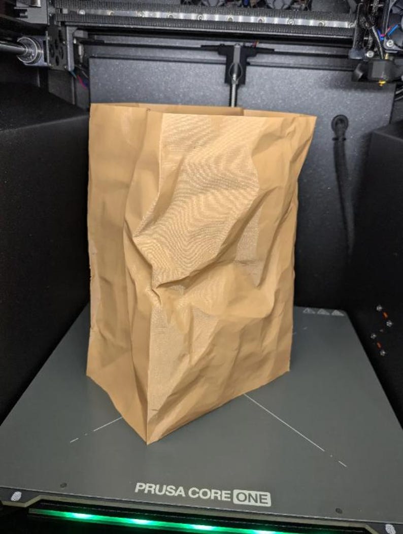 May include: A crumpled brown paper bag stands upright on a gray surface, likely inside a 3D printer. The bag has a rectangular base and an open top, with visible creases and folds throughout its surface. The printer's components are visible in the background.