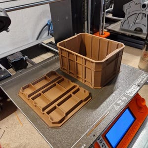 Könnte beinhalten: Ein brauner Aufbewahrungskorb und sein Deckel liegen auf dem Druckbett eines 3D-Druckers. Der Korb hat ein gitterartiges Design, und der Deckel weist rechteckige Fächer auf. Der Drucker ist orange und schwarz, mit einem digitalen Display. Die Szene deutet auf 3D-gedruckte Aufbewahrungslösungen hin.