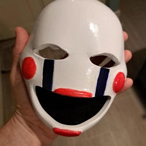 以下が含まれることがあります： 赤と黒のアクセントが付いた白いマスク。マスクには頬に赤い円、青い縦線、赤い笑顔があります。目の穴は楕円形です。このマスクは小道具またはコスチュームの一部です。
