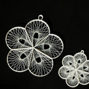 Puede incluir: Dos adornos blancos con forma de flor con intrincados diseños de arte de hilo. El adorno más grande tiene cinco pétalos, mientras que el más pequeño tiene seis. Ambos tienen un lazo para colgar y están sobre un fondo negro.