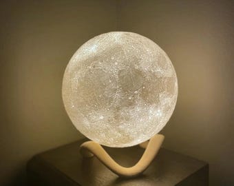 May include: A glowing, spherical moon lamp with a textured surface, resting on a cream-colored, curved stand. The lamp is placed on a dark brown nightstand, creating a warm, ambient light. The lamp's diameter is approximately 8 inches.