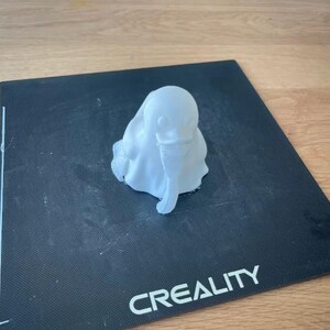 May include: A small, white 3D-printed sculpture of a ghost-like figure with a rounded head and draped body. The sculpture is sitting on a black surface with the word "CREALITY" printed in white.