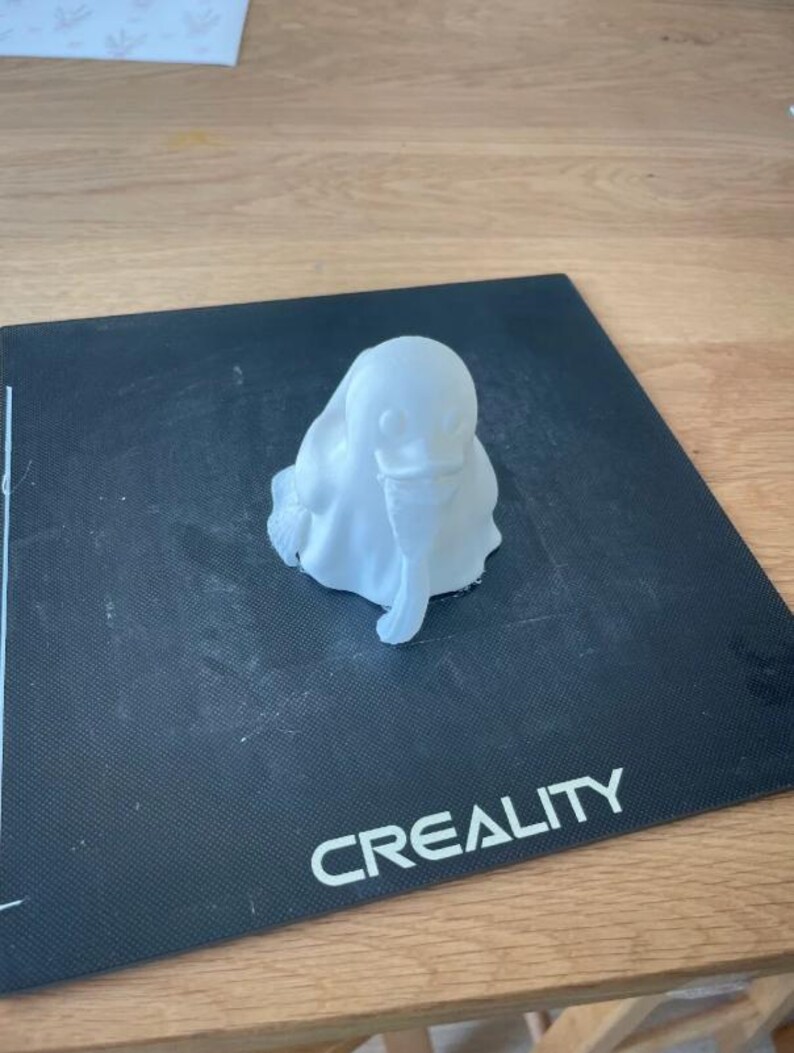 May include: A small, white 3D-printed sculpture of a ghost-like figure with a rounded head and draped body. The sculpture is sitting on a black surface with the word "CREALITY" printed in white.