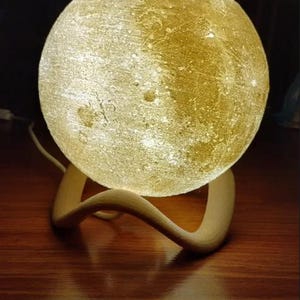 May include: A glowing, spherical moon lamp with a textured surface, resting on a curved wooden base. The lamp emits a warm, soft light, creating a cozy ambiance. The lamp is a decorative home decor item.