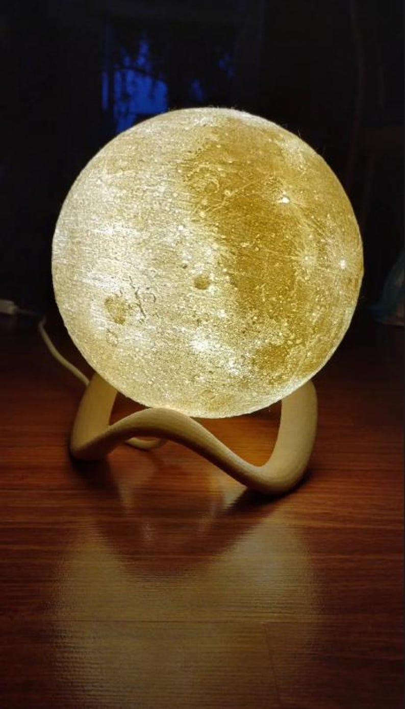 May include: A glowing, spherical moon lamp with a textured surface, resting on a curved wooden base. The lamp emits a warm, soft light, creating a cozy ambiance. The lamp is a decorative home decor item.