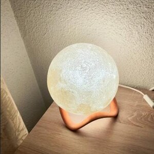 May include: A glowing, spherical moon lamp with a textured surface, resting on a curved, orange-colored base. The lamp is placed on a wooden nightstand, with a cord extending to the right.