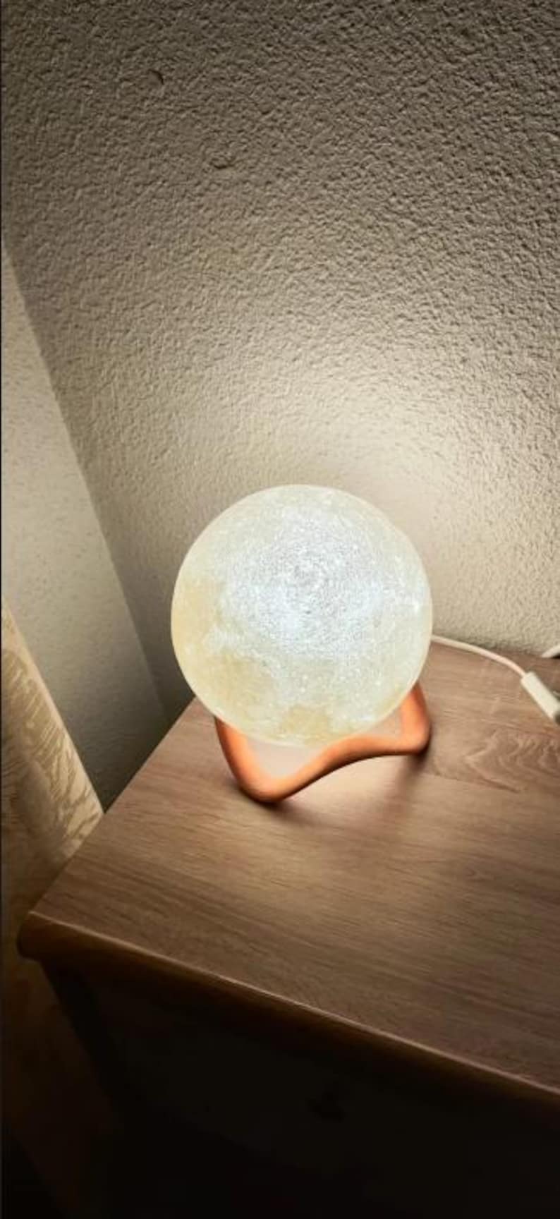May include: A glowing, spherical moon lamp with a textured surface, resting on a curved, orange-colored base. The lamp is placed on a wooden nightstand, with a cord extending to the right.