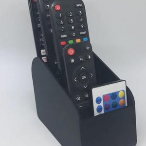 May include: A black remote control tidy containing several black remote controls and a small white remote. The organiser is made of a matte black material and has a curved design. The remotes have various buttons and numbers.
