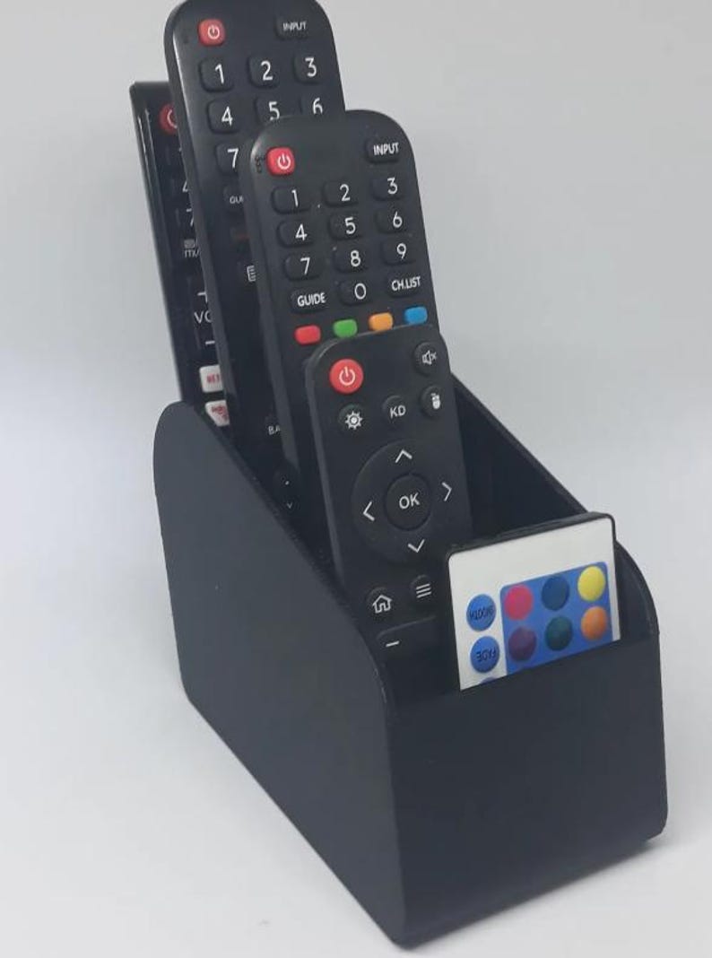 May include: A black remote control tidy containing several black remote controls and a small white remote. The organiser is made of a matte black material and has a curved design. The remotes have various buttons and numbers.