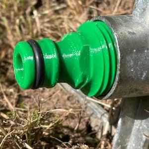 Puede incluir: Primer plano de un conector de manguera de plástico verde con una junta tórica negra, fijado a un accesorio metálico. El conector tiene un diseño acanalado, sobre un fondo borroso de hierba seca y tierra.