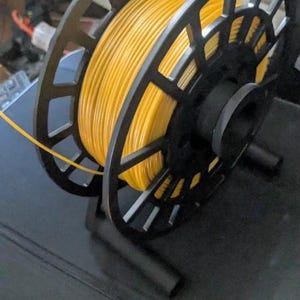 Puede incluir: Un soporte negro para bobinas de filamento de impresora 3D con una bobina de filamento amarillo. El soporte tiene un dise&ntilde;o geom&eacute;trico y est&aacute; hecho de pl&aacute;stico negro. El filamento amarillo est&aacute; enrollado alrededor de la bobina.