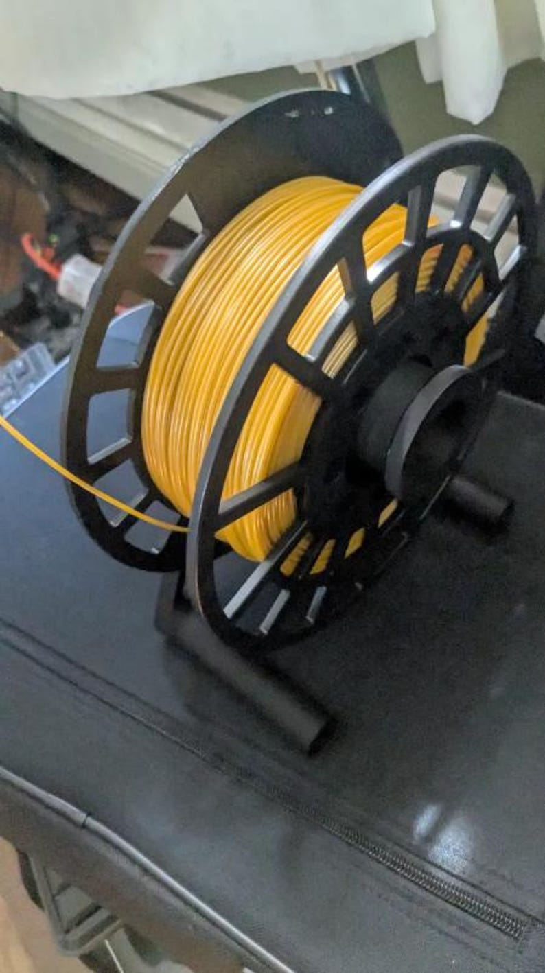 Puede incluir: Un soporte negro para bobinas de filamento de impresora 3D con una bobina de filamento amarillo. El soporte tiene un dise&ntilde;o geom&eacute;trico y est&aacute; hecho de pl&aacute;stico negro. El filamento amarillo est&aacute; enrollado alrededor de la bobina.