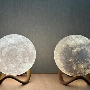 May include: Two moon-shaped lamps with detailed surface textures, each resting on a curved, wooden base. One lamp emits a bright white light, while the other glows with a softer, warmer tone. The lamps are designed to resemble the moon.