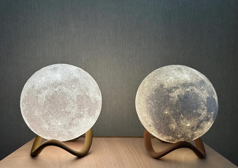 May include: Two moon-shaped lamps with detailed surface textures, each resting on a curved, wooden base. One lamp emits a bright white light, while the other glows with a softer, warmer tone. The lamps are designed to resemble the moon.