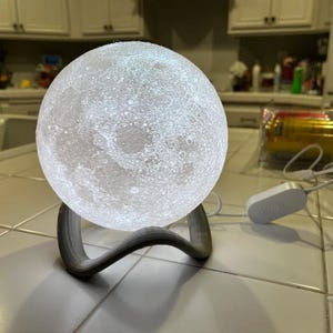 May include: A glowing white moon-shaped lamp with detailed surface features, resting on a gray, curved stand. The lamp is illuminated, showcasing craters and textures. A white power cord and adapter are connected to the lamp.