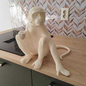 Może przedstawiać: Kremowa rzeźba małpy w pozycji siedzącej, trzymająca mały kubek. Rzeźba stoi na drewnianym blacie z czarną płytą grzejną i wzorzystą osłoną z płytek. Ogon małpy rozciąga się w prawo.