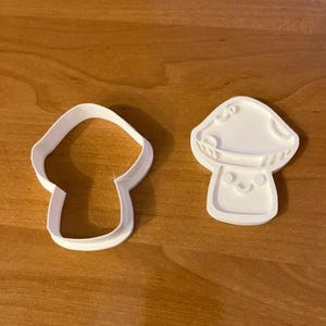 May include: Two white mushroom-shaped cookie cutters. One is a simple outline, and the other has a detailed mushroom design with a smiling face. The cutters are made of plastic and sit on a wooden surface.
