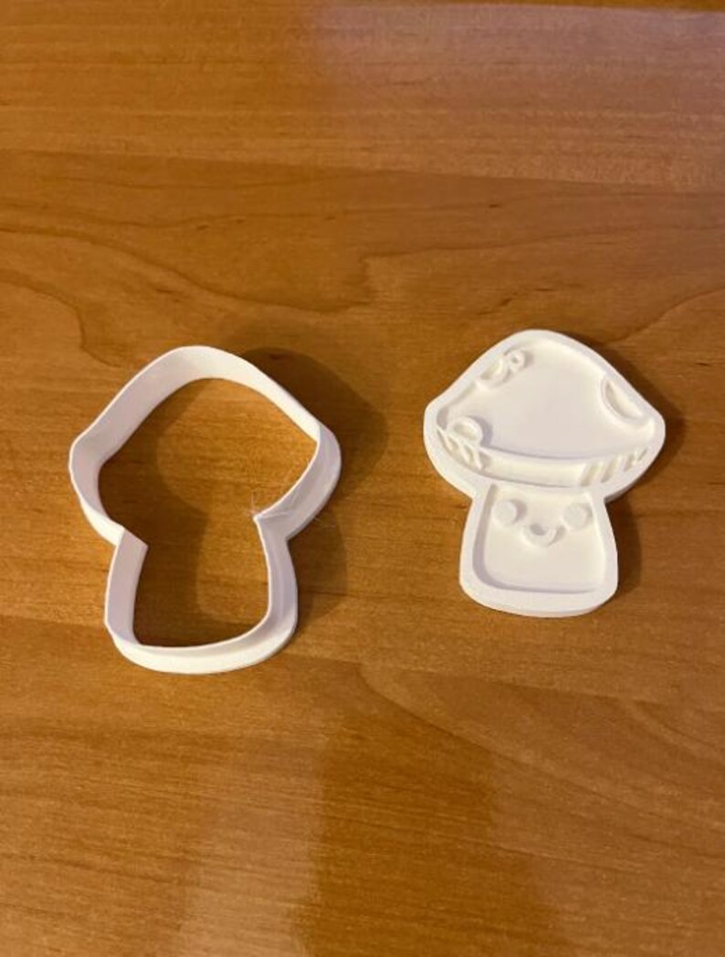 May include: Two white mushroom-shaped cookie cutters. One is a simple outline, and the other has a detailed mushroom design with a smiling face. The cutters are made of plastic and sit on a wooden surface.