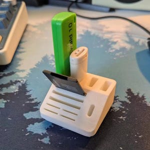 May include: A white 3D-printed organiser with slots for electronic accessories. A green USB drive with the text "Ewald 10" is visible, along with a white adapter and a black memory card. The organiser is on a patterned surface.