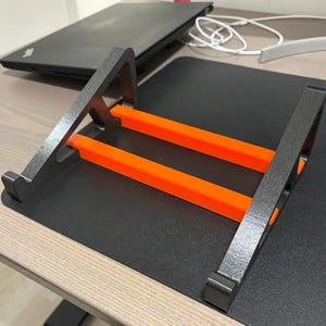 May include: A black laptop stand with two orange support bars. The stand is on a black surface, with a laptop and white cable in the background. The stand is designed to elevate a laptop for better ergonomics.