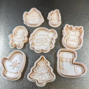 Op de afbeelding: Een verzameling van acht beige plastic kerstkoekjessnijders. De vormen omvatten een kerstman, een rendier, een kerstboom, een kous, een want, een peperkoekmannetje en andere feestelijke ontwerpen.