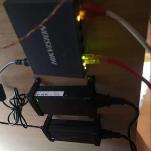 Könnte beinhalten: Schwarzer HIKVISION-Netzwerk-Switch mit mehreren Anschlüssen, verbunden mit roten und weißen Ethernet-Kabeln. Zwei schwarze Netzadapter sind darunter montiert. Der Switch und die Adapter befinden sich auf einer Holzoberfläche.