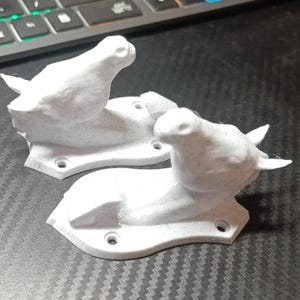 Op de afbeelding: Twee witte, vogelvormige wandhaken. Elke haak heeft een vogel die op een basis met drie schroefgaten zit. De haken zijn gemaakt van een textuurrijk materiaal en zijn ontworpen voor wandmontage.
