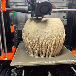 May include: A beige, textured, spherical object with finger-like protrusions is being 3D printed. The printer is an Original Prusa i3, with orange and black components. The print bed is visible, along with the printer's nozzle and cooling fan.