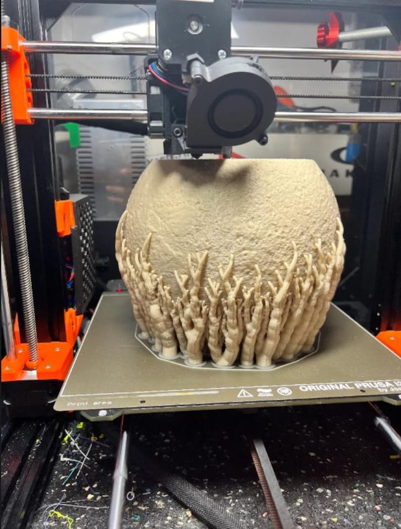 May include: A beige, textured, spherical object with finger-like protrusions is being 3D printed. The printer is an Original Prusa i3, with orange and black components. The print bed is visible, along with the printer's nozzle and cooling fan.