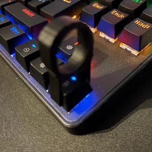 Könnte beinhalten: Ein schwarzes ringförmiges Objekt ist an einer schwarzen mechanischen Tastatur befestigt. Die Tastatur hat schwarze Tasten mit mehrfarbiger Hintergrundbeleuchtung. Der Ring befindet sich über den Tasten, mit Fokus auf der Ecke der Tastatur.