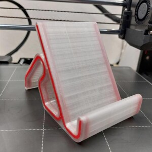 May include: A 3D-printed phone stand with a clear body and red trim. The stand is angled for optimal viewing and has a curved base to hold a phone securely. The design is modern and functional, suitable for desks or tables.