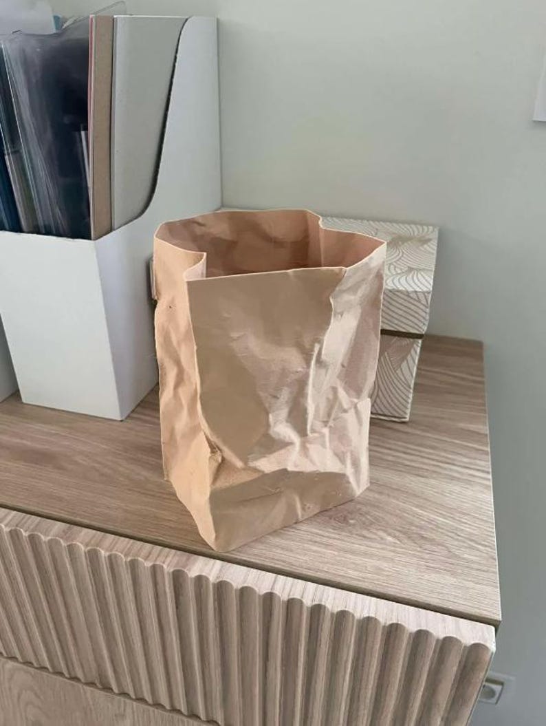 May include: A crumpled, open-top paper bag in a light brown hue sits on a wooden surface. The bag has a rectangular base and a textured appearance. A white organizer and a decorative box are in the background.