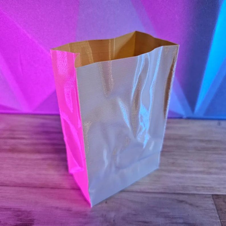 May include: A rectangular paper bag with a metallic silver front and a bright pink side. The inside and top edge of the bag are a golden color. The bag is empty and stands upright.