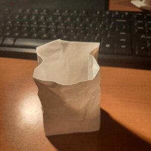 May include: A small, open, white paper bag stands upright on a wooden surface. The bag has a rectangular base and a slightly crumpled appearance. The lighting creates a shadow to the right of the bag.