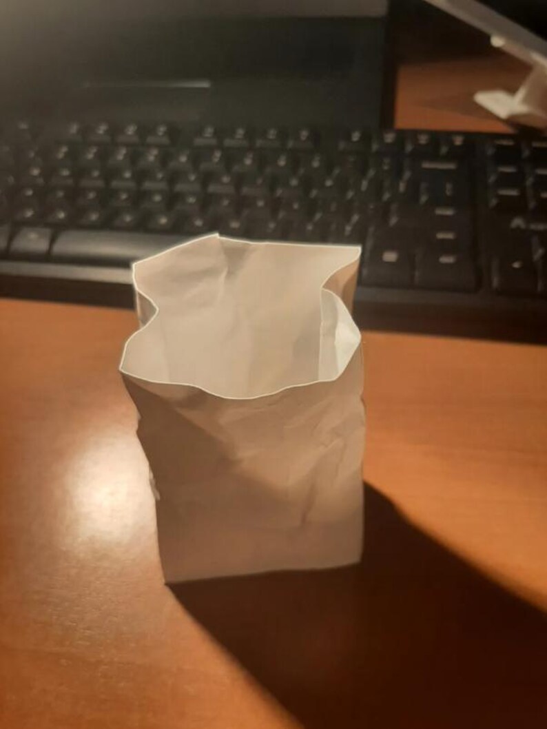 May include: A small, open, white paper bag stands upright on a wooden surface. The bag has a rectangular base and a slightly crumpled appearance. The lighting creates a shadow to the right of the bag.