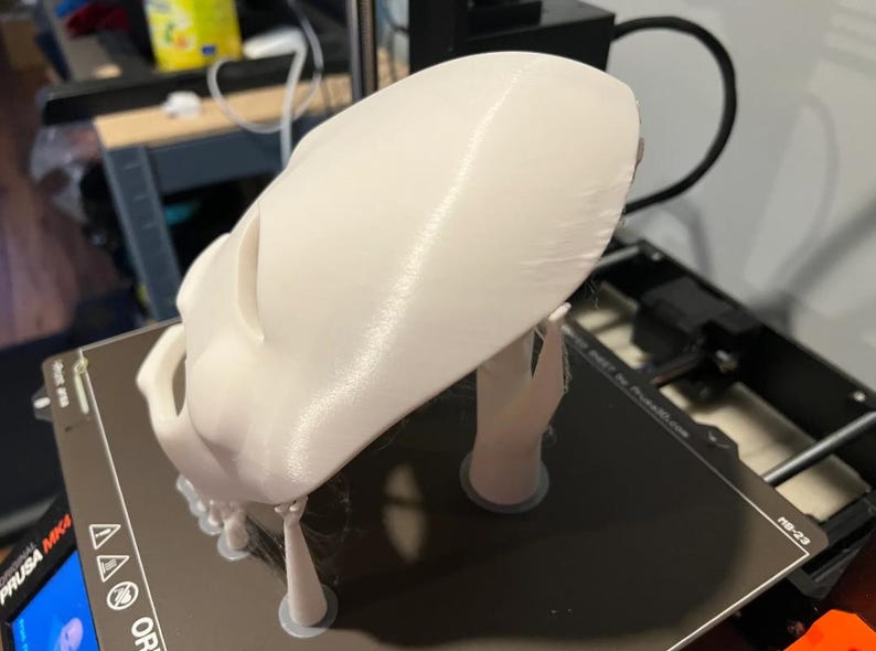 以下が含まれることがあります： 白の3Dプリントされた頭蓋骨モデル。おそらく試作品または装飾品。頭蓋骨は、おそらくプリンターベッドである黒い表面に置かれており、まだサポート構造が取り付けられています。モデルは詳細で、印刷されたばかりのようです。