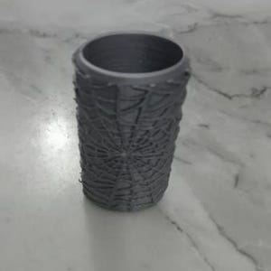 May include: A gray cylindrical container with a textured, web-like pattern. The container is made of a solid material and has a smooth, open top. The design gives the container a unique, artistic appearance.