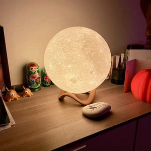 May include: A glowing, spherical moon lamp with a textured surface, resting on a curved wooden stand. The lamp emits a soft, warm light. Additional decorative items include Russian nesting dolls and small orange alien figurines.