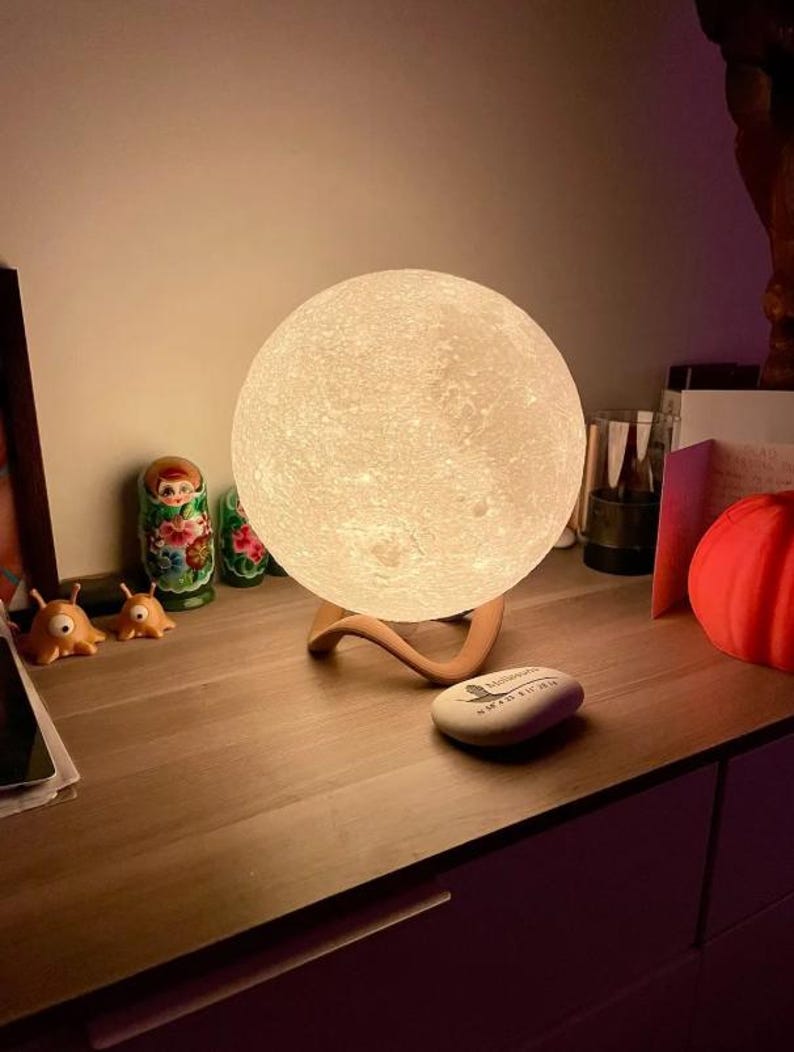 May include: A glowing, spherical moon lamp with a textured surface, resting on a curved wooden stand. The lamp emits a soft, warm light. Additional decorative items include Russian nesting dolls and small orange alien figurines.
