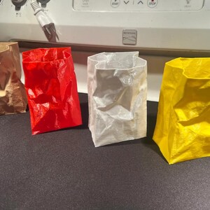 May include: Four colorful paper bags in brown, red, white, and yellow. The bags are crumpled and have an open top. They are arranged in a row on a dark surface, with a white appliance in the background.