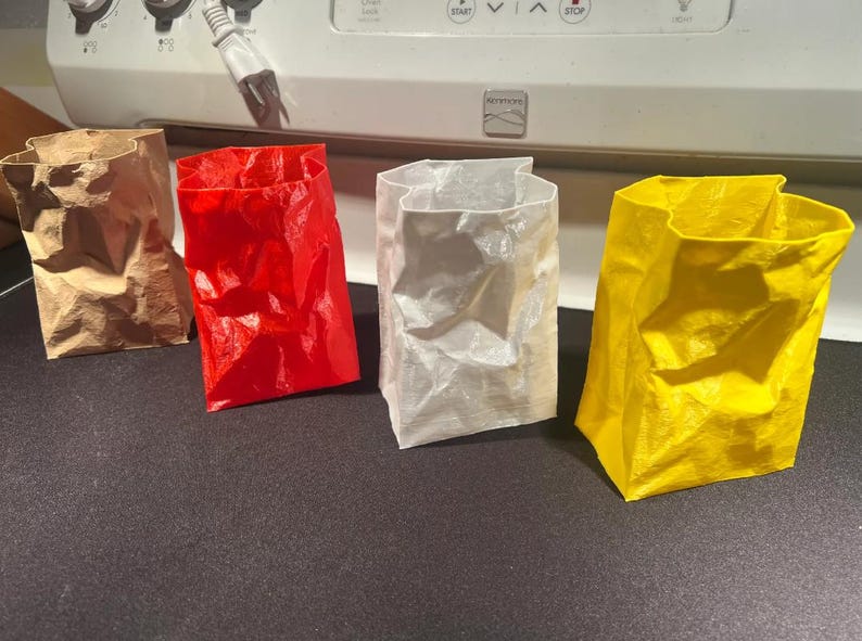 May include: Four colorful paper bags in brown, red, white, and yellow. The bags are crumpled and have an open top. They are arranged in a row on a dark surface, with a white appliance in the background.
