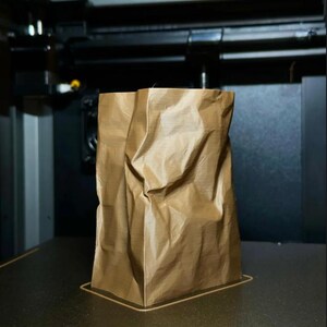 May include: A brown, crumpled paper bag stands upright on a dark surface. The bag has a rectangular base and is slightly creased, suggesting it is made of a flexible material. The background shows a glimpse of machinery.