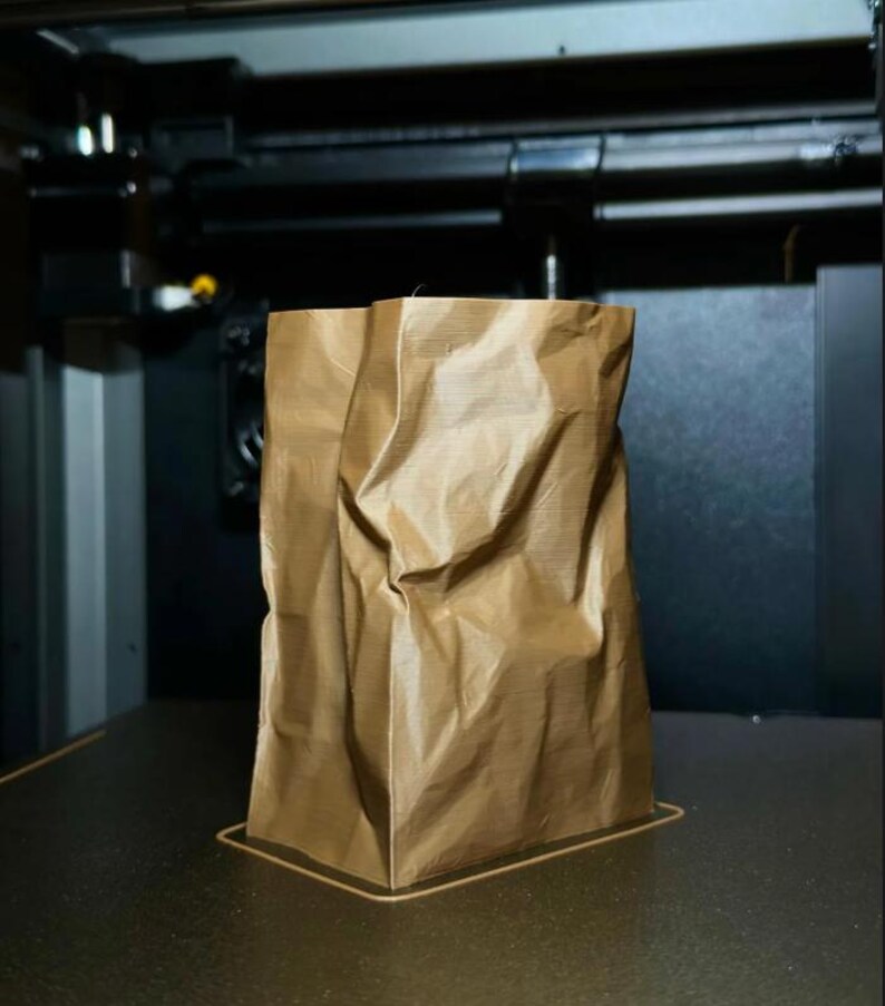 May include: A brown, crumpled paper bag stands upright on a dark surface. The bag has a rectangular base and is slightly creased, suggesting it is made of a flexible material. The background shows a glimpse of machinery.
