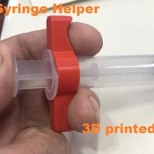 Puede incluir: Una herramienta de ayuda para jeringas roja impresa en 3D está unida a una jeringa transparente. Los textos "Syringe Helper" y "3D printed" son visibles en naranja. La jeringa tiene marcas negras.