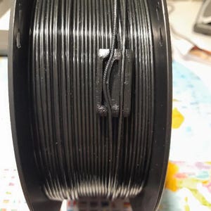May include: A black spool of 3D printer filament. The filament is tightly wound around the spool, and a black clip secures the end. The spool is made of black plastic and is ready for use in a 3D printer.