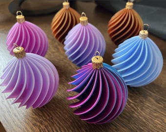 May include: A collection of colorful, spiral-designed Christmas ornaments. The ornaments are in shades of pink, purple, blue, and bronze, each with a gold-colored cap for hanging. The design features a series of curved, layered segments.
